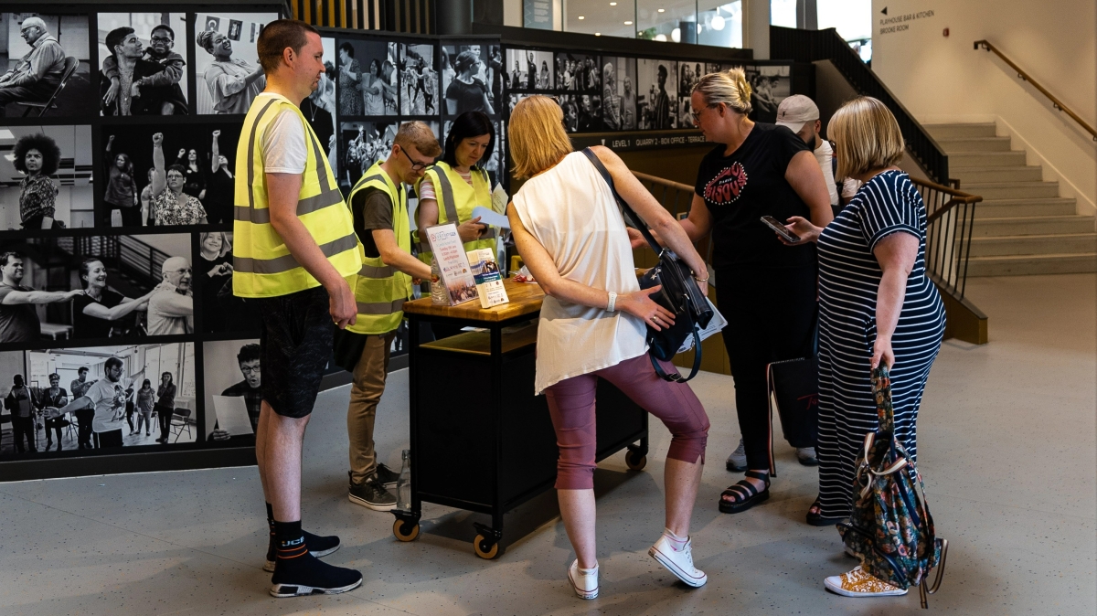 An image of volunteers wearing hi-visibility vests, welcoming people to the 2023 AutistiCon event. They are stood at a table with wheels on the bottom of each leg.